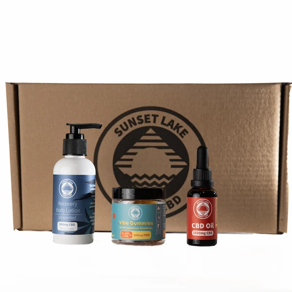 A cardboard box behind three Sunset Lake CBD products. From left to right we have the Unscented CBD Recovery Body Lotion, the Full Spectrum CBD Vibe Gummies, and the 1500mg Full Spectrum CBD Oil Tincture