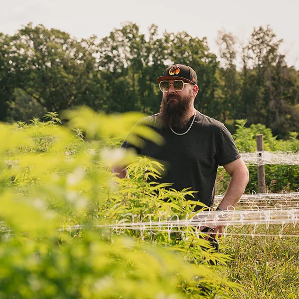 Rev standing in the field at Sunset Lake CBD's Vermont hemp farm. He's got a big beard and is wearing large white sunglasses and a black baseball hat.