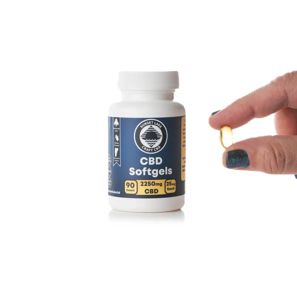 Two fingers pinching a CBD Softgel next to a bottle of softgel capsules