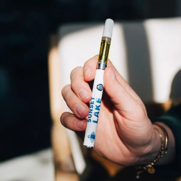 A hand holding a CBD vape cart attached to a branded Sunset Lake Vape Battery